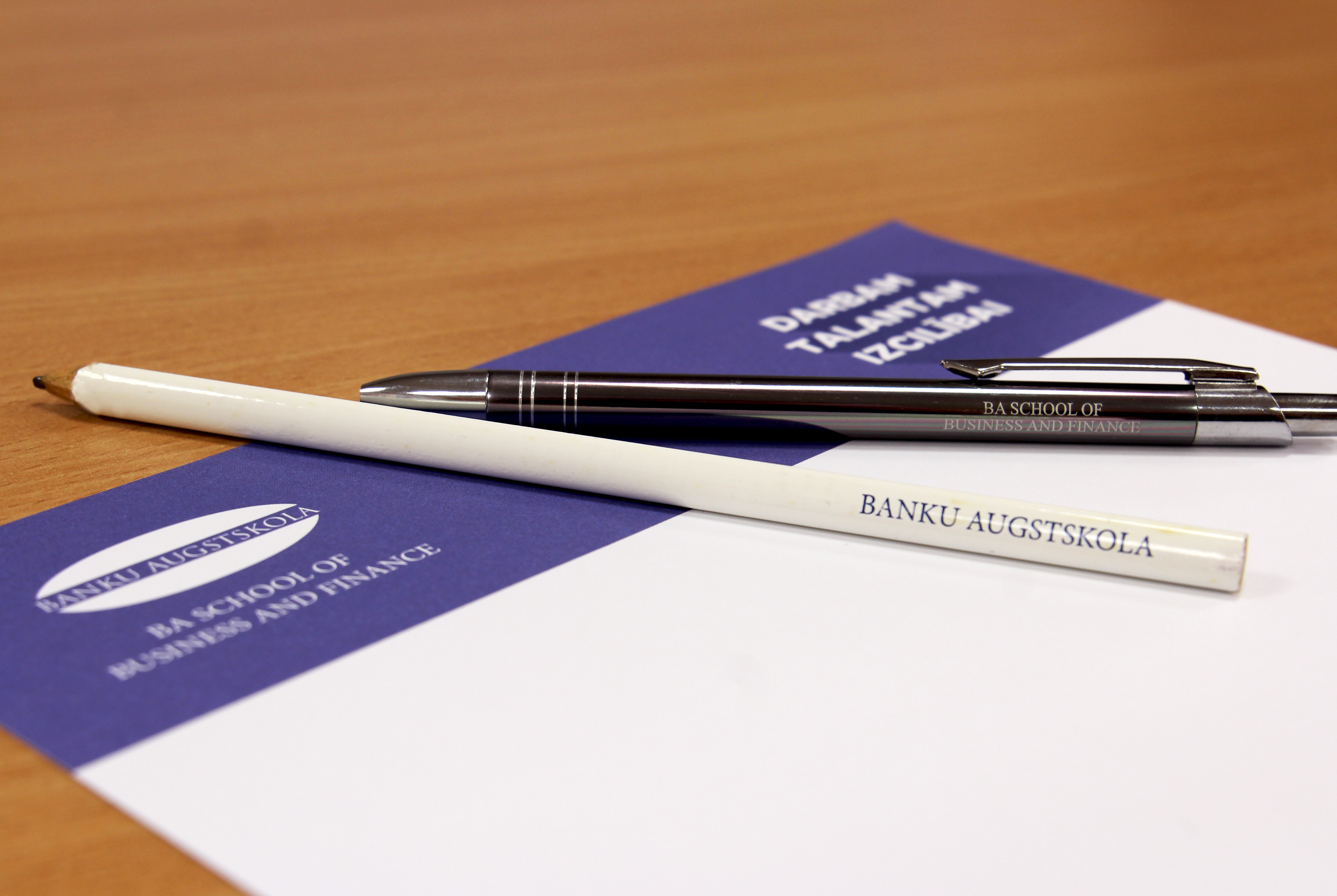 Galda tuvplāns ar divām pildspalvām un piezīmju blociņu ar LU Banku augstskolas logo.